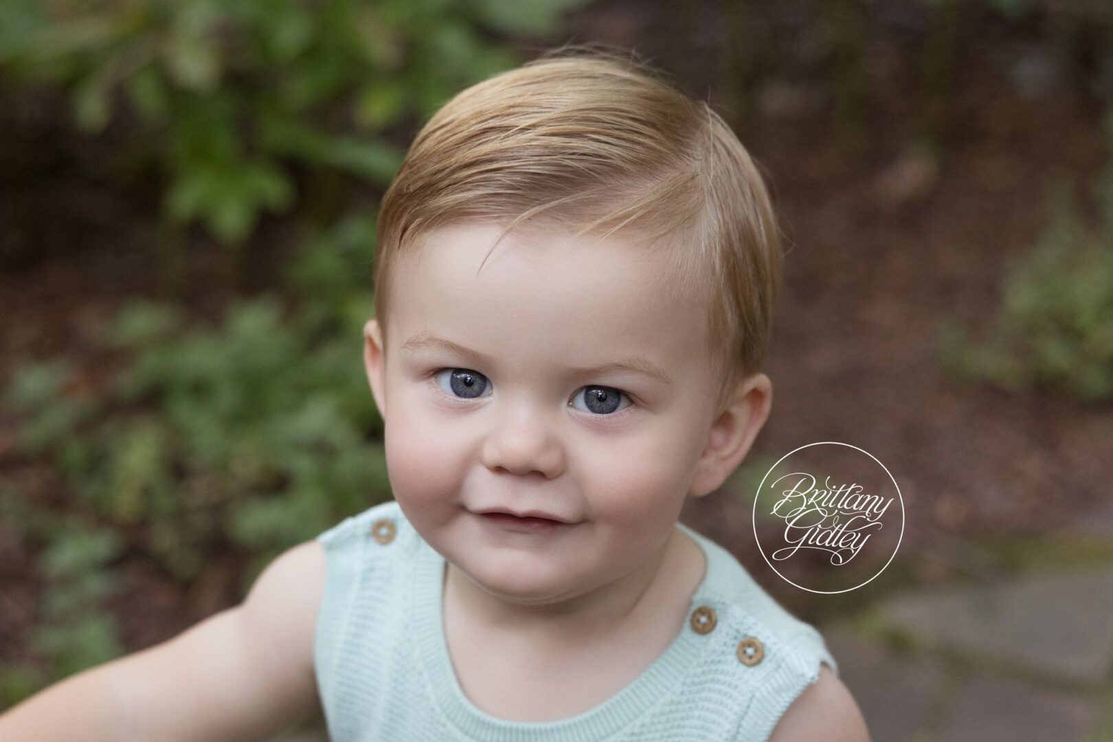 Close-up of a smiling one year old boy in a mint romper in the gardens at Stan Hywet