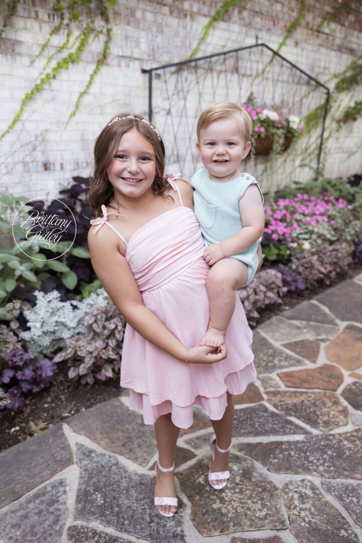 Big sister in a pink dress holding her baby brother on a stone path at Stan Hywet Hall