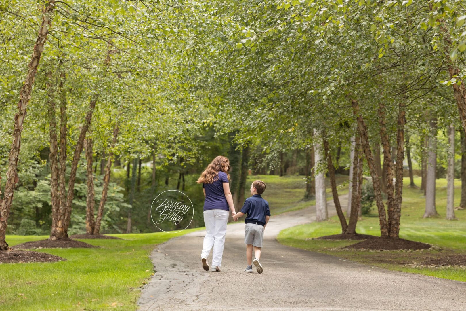 Mom and son walking hand in hand down a tree-lined drive during an in-home family session