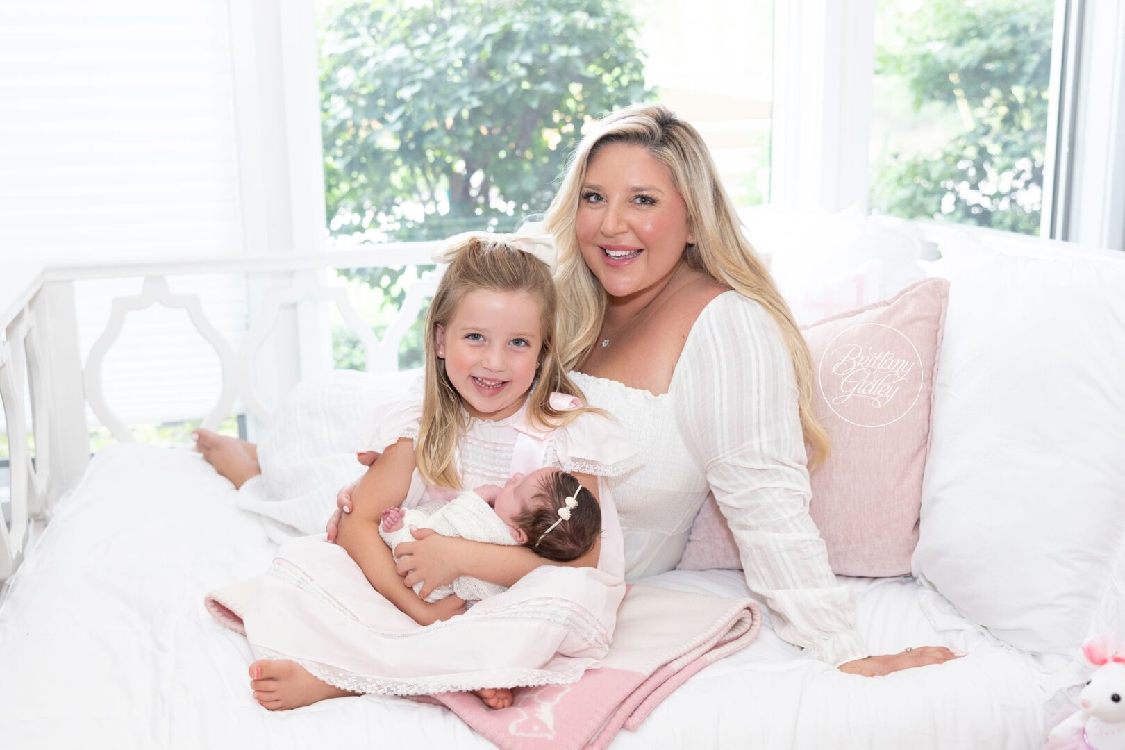 Big sister cuddling her newborn sister with Mom on a white bed in Naples, Florida