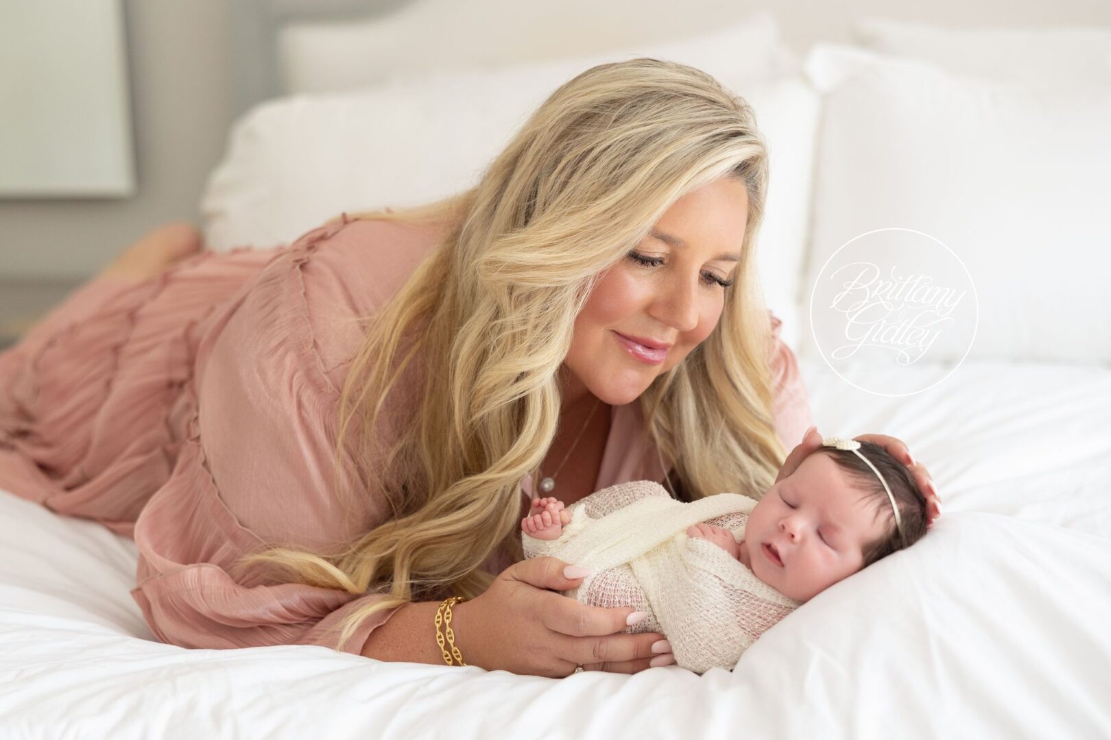 Mom in a blush dress snuggling her newborn daughter on a white bed