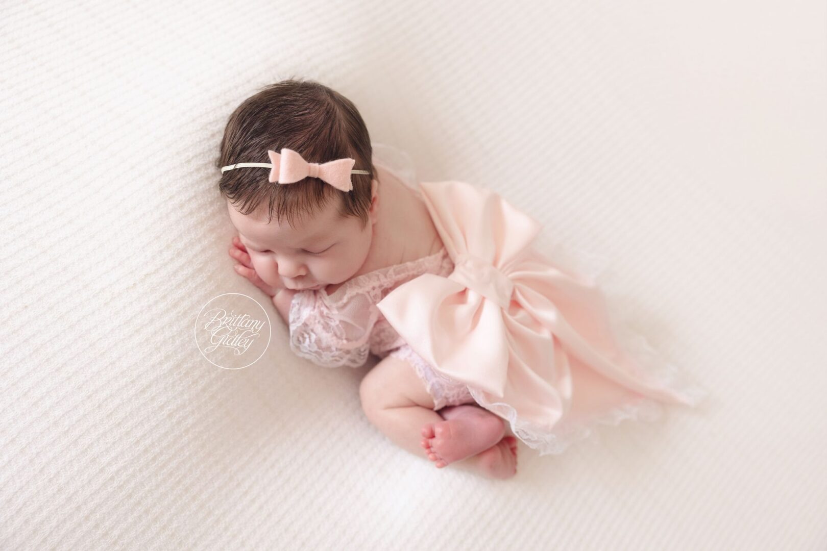 Newborn baby girl in a pink bow outfit asleep on a cream blanket in Naples, Florida