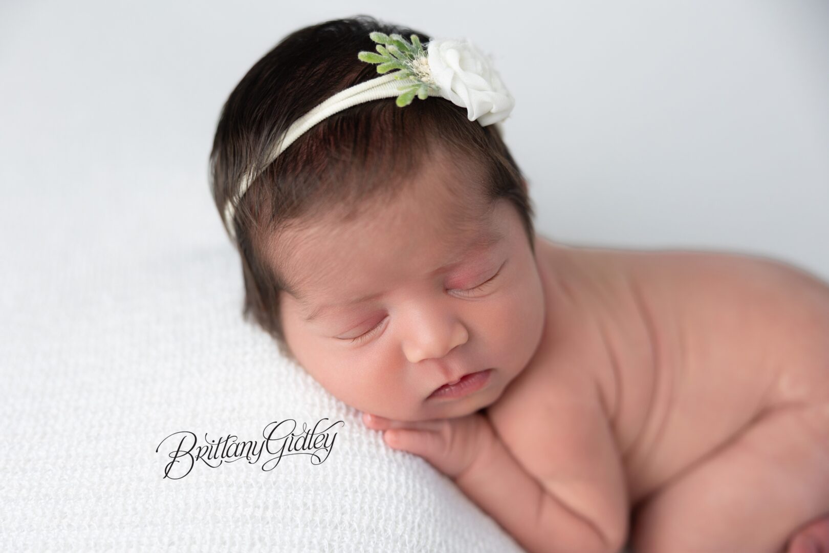 Newborn Ava resting her cheek on her hands with a flower headband on white