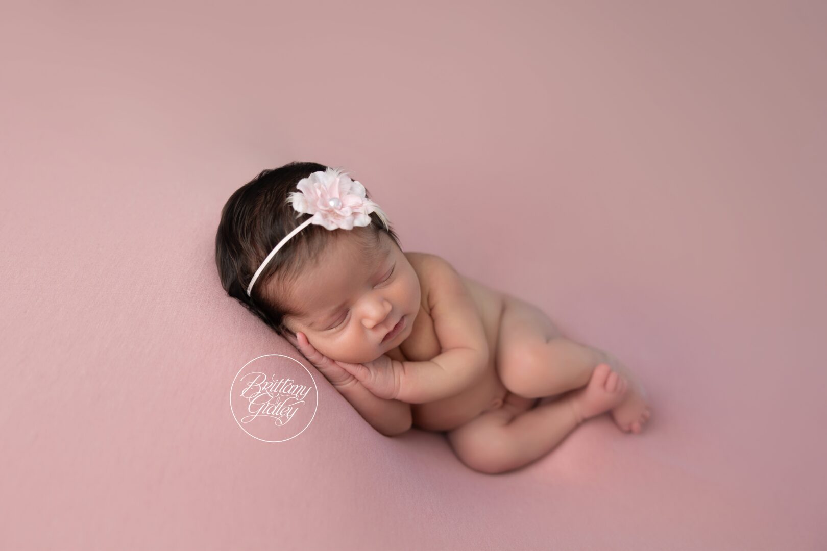 Newborn Ava curled up asleep on soft pink with a flower headband